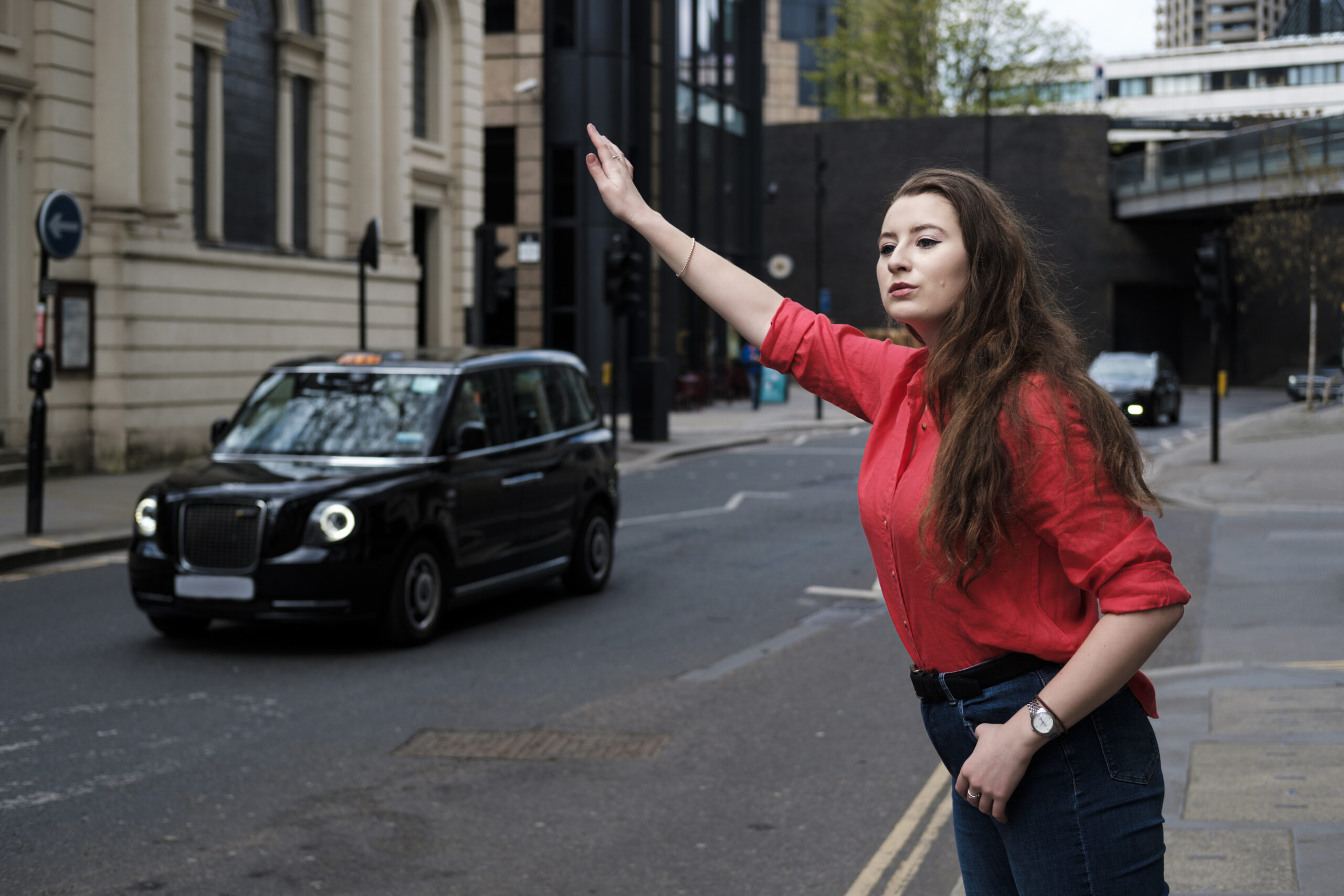 Young woman in red shirt hailing a taxi in urban London. Gesturing to signal to a cab driver.